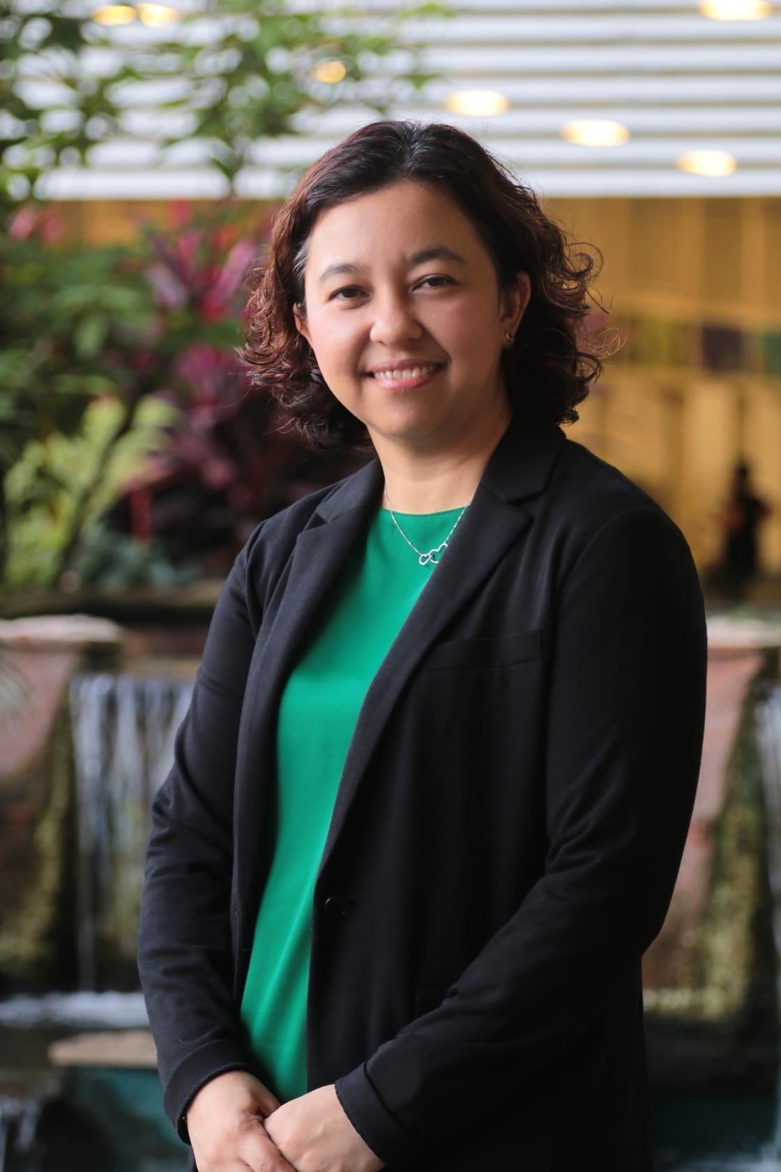 A smiling woman with curly dark hair wears a black blazer and green top, standing before an indoor fountain.